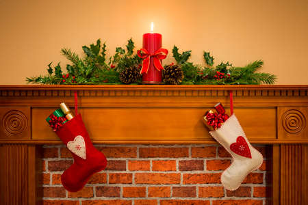 Two Christmas stockings with gift wrapped presents hanging on a mantelpiece over a fireplace, plus burning candle with festive garland including holly, fir and pine cones.の写真素材