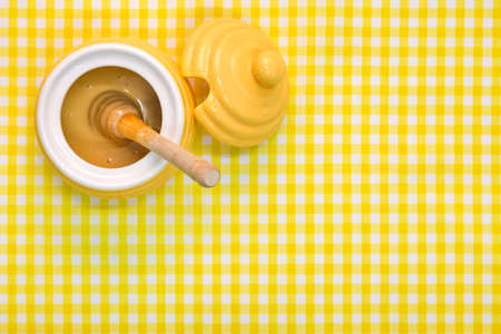 A beehive themed pot full of Manuka honey with wooden dipper on a yellow gingham background with copy space.の写真素材