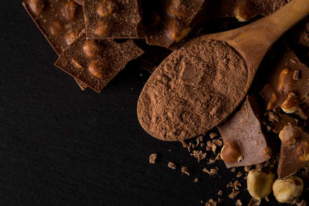 Broken chocolate nuts pieces and cocoa powder in spoon on dark stone background