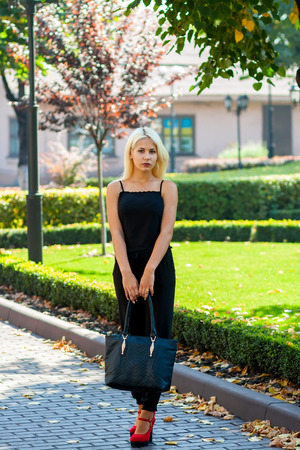 Young beautiful blonde girl posing on the background of the urban landscape. Sexy lady in a black dress and red shoes with a pleasant appearance.の写真素材