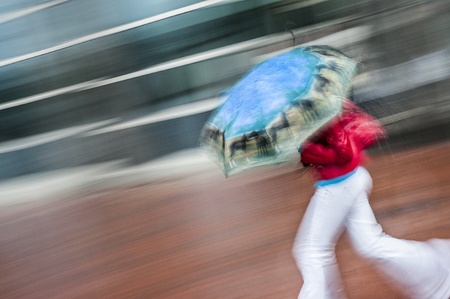 Female running from rain in red coat and white pants with blue umbrellaの写真素材