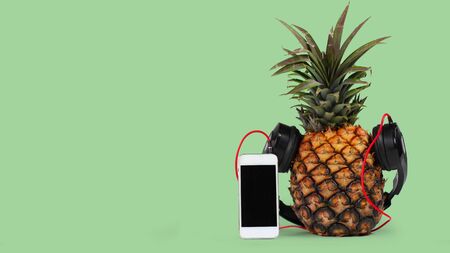 fresh pineapple with black headphones and smartphone with isolated black screen against green backgroundの写真素材