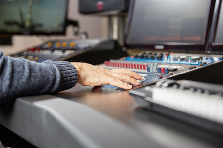 Hands of a man on a production consoe with a keyboard in front and monitors in the back