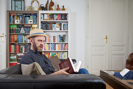 Hipster with hat reading a book at home in an old building with a book shelf in the backgroundの写真素材