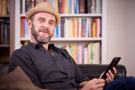 Hipster with hat sitting on a couch in his liviing room with a mobile phoneの写真素材