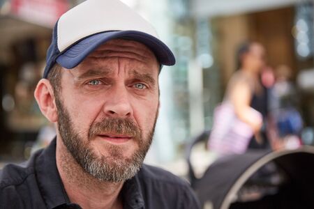 Hipster with beard and baseball cap sitting in a street cafe looking seriously