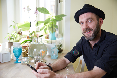 Hipster with hat and beard sitting happy in his kitchen at home looking at phone and soap bubbles in the airの写真素材