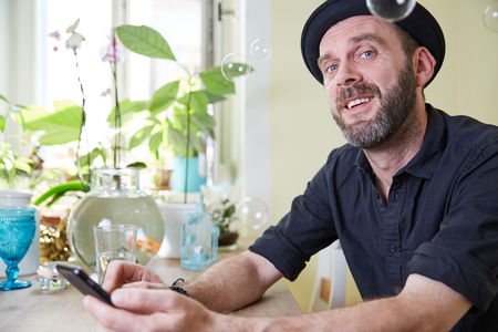 Hipster with hat and beard sitting happy in his kitchen at home looking at phone and soap bubbles in the airの写真素材