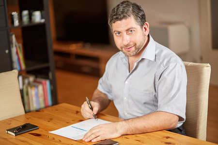 Caucasian man smiling and writing contract document sitting on table at homeの写真素材