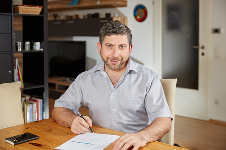 Caucasian man smiling and writing contract document sitting on table at homeの写真素材