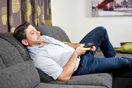 Caucasian man sitting on couch at home watching tv with remote control in his handの写真素材