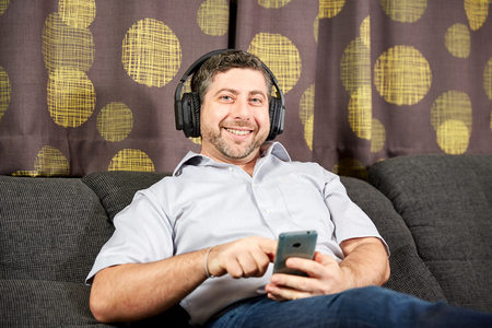 Man with headphones and phone smiling on sofa at homeの写真素材