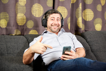 Man with headphones pointig at phone on sofa at home
