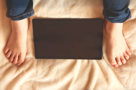 Teenager sitting on the bed and looking at tablet screen. Tablet between legs. Home education....
