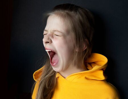 Teenager girl in an orange sweatshirt with bright emotions on her face on a black background. Girl screaming. Selective focus. Side viewの写真素材