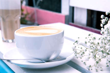 Cappuccino in a white ceramic cup and bouquet of small white flowers on a wooden bench. Open-air rustic morning coffee. Morning routine. Gypsophila flowers. Side viewの写真素材