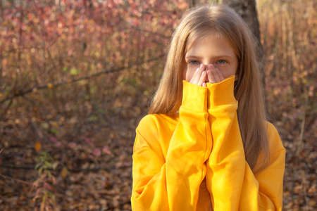 Beautiful teen blonde girl in a bright orange sweatshirt covers her face with her hands in an autumn park. Autumn portrait in the forest against the background of fallen golden leavesの写真素材