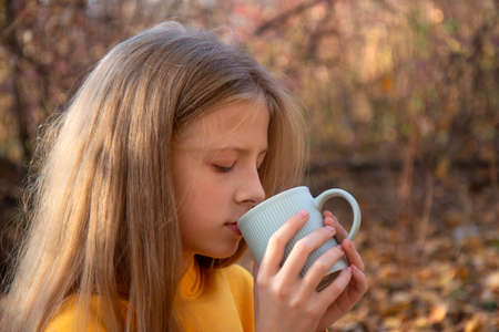 Beautiful teen blonde girl in a bright orange sweatshirt with a cup of tea in an autumn park. Autumn...