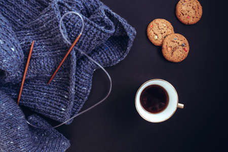 Blue knit sweater, knitting needles, porcelain gold cup with coffee and flavored cookies with colored drops on black background. Knitwear as a concept of female hobbyの写真素材