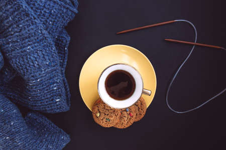 Blue knit sweater, knitting needles, porcelain gold cup with coffee and yellow plate with flavored cookies with colored drops on black background. Knitwear as a concept of female hobbyの写真素材