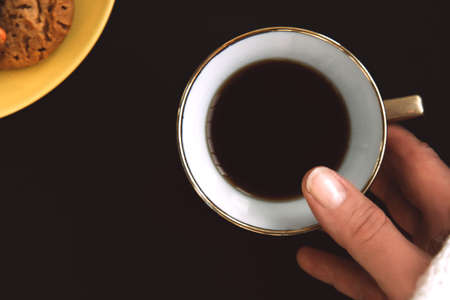 Woman's hand is holding a golden porcelain cup of coffee on black background. Yellow plate with flavored cookies with colored dropsの写真素材