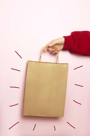 Female hand in a red sweater holds a craft bag for shopping on a bright pink background. Ecological...