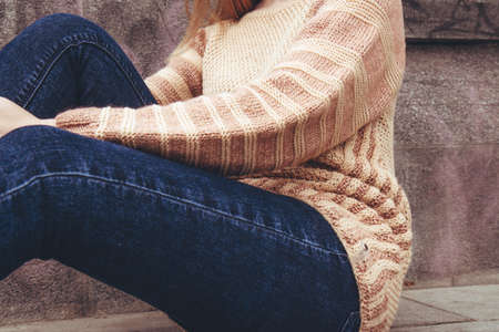 The girl is sitting on the sidewalk. Young beautiful woman in a cozy beige, knitted sweater spends...