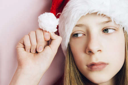 Smiling beautiful girl with long blonde hair in fluffy Santa Claus hat on a pink background. Sad teenage girl holding a bubon on her hatの写真素材