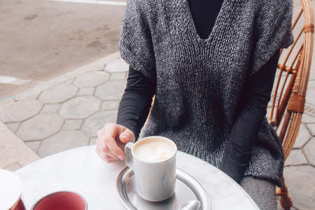 Woman in a cozy gray-blue vest spends time outdoors in the city. Female sits in street cafe on a wooden wicker chair in street cafe. Cup of morning coffee. Nofaceの写真素材