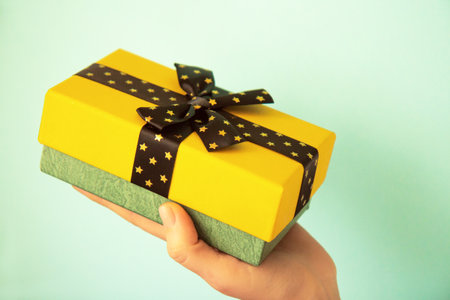 Yellow gift box with black bow with golden stars in woman hand on light green backgroundの写真素材