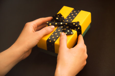 Yellow gift box with black bow with golden stars in woman's hands on a dark background