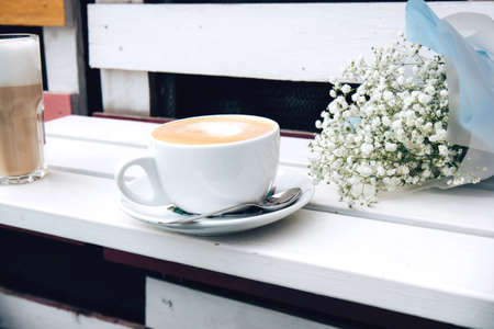 Cappuccino in a white ceramic cup and a latte in a glass with a straw on a white wooden bench. Open-air rustic morning coffeeの写真素材