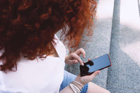 One woman with bright curly red hair on the street looks at her mobile phone. Woman outside the house. Casual life of a female. Lifestyleの写真素材