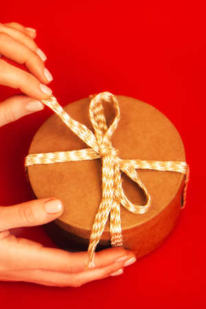 Craft gift box with beige rope in woman's hands on a bright red background. Top view