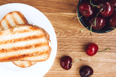 Grilled toast sandwich with egg on a white plate and a large, ripe, red cherry in a white cup on a wooden background. Toasted breads for breakfast. Light and quick breakfastの写真素材