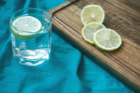 Slices of yellow lemon lie on wooden cutting board. Glass of water with slice of lemon on a background of turquoise fabric. Tropical fruits. Fresh citrus. Top viewの写真素材