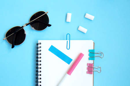 White open notebook with blue paper clip, clip and piece of scotch tape, gum and sunglasses on blue...