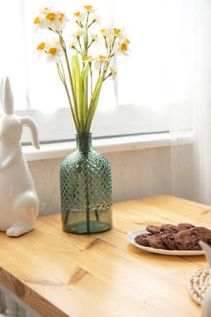Narcissus flowers in a beautiful vase, chocolate cookies on a plate on a wooden table by the window....