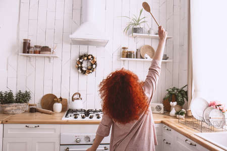 Cheerful happy woman with bright curly red hair dancing on the white, light kitchen in the morningの写真素材