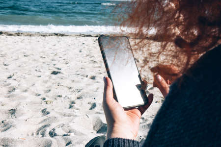 One woman with bright curly red hair by the ocean looks at her mobile phone. Woman outside the house. Casual life of a female. Lifestyleの写真素材