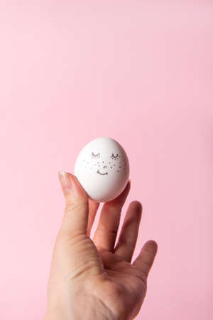 Easter holiday concept with cute handmade egg. Cute face egg in a female hand on a pink background