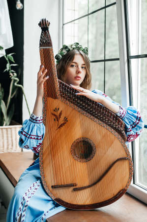 A girl in traditional Ukrainian clothes, with a bandura in her hands, sits on the windowsillの写真素材
