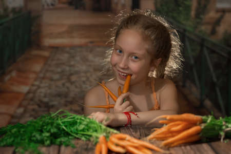 A girl in an orange sundress is sitting at a wooden table with a young carrot on it. The girl holds a carrot in her hands and nibbles it. The girl's face is illuminated by the sunlight.の写真素材