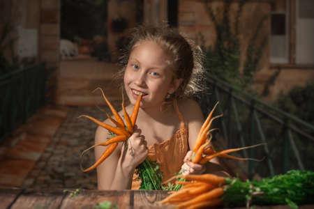 A girl in an orange sundress is sitting at a wooden table with a young carrot on it. The girl holds a carrot in her hands and nibbles it. The girl's face is illuminated by the sunlight.の写真素材