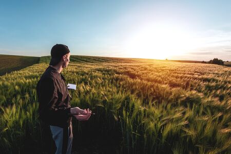 Young agronomist in cap takes notes in a notebook on a green agricultural field during sunset