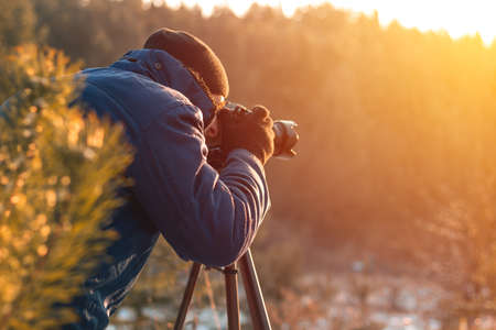 A man in warm clothes in winter makes beautiful shots with a digital camera on a tripod, a portrait at sunset, winter photosの写真素材