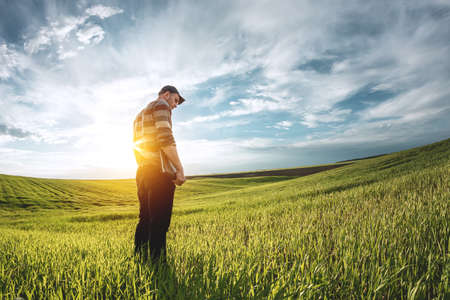 A young agronomist holds a folder in his hands on a green wheat field. A farmer makes notes on the background of agricultural land during sunset. Man in a cap with a folder of documentsの写真素材