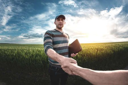A young agronomist holds a folder in his hands on a green wheat field. A farmer makes notes on the background of agricultural land during sunset. Man in a cap with a folder of documentsの写真素材
