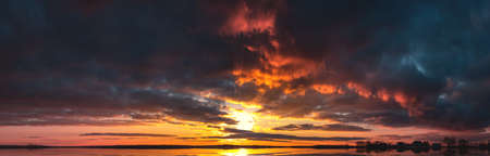 A bright sunset with a large yellow sun reflected in the water. Sunrise at the edge of the village. The big sun goes over the horizon reflecting the light in the lakeの写真素材