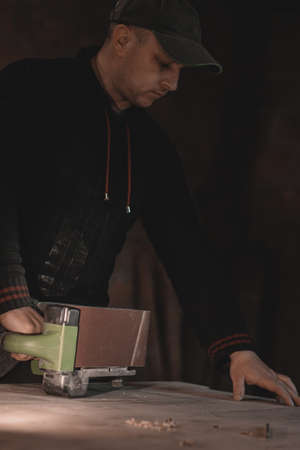A carpenter works in a workshop. Joiner's grinders, furniture manufacturing. A carpenter is grinding a wooden part with an electric sander.の写真素材